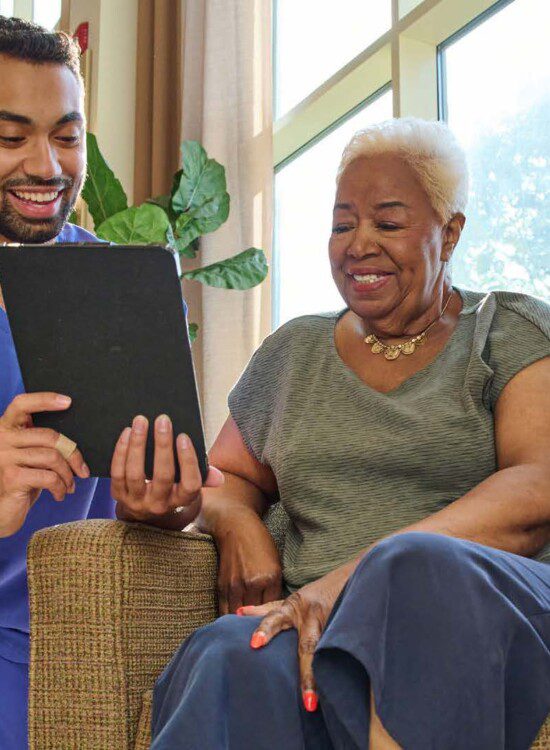 seated senior woman smiles at a tablet, held by her kneeling caregiver